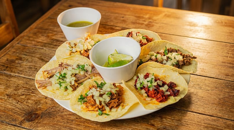 A white plate with various tacos, lime wedges, and a bowl of green salsa on a wooden table.